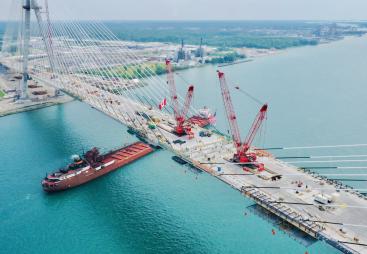 Aerial view of a bridge under construction across a river with two red cranes on its deck