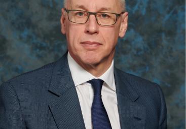 A studio head and shoulders photo of a man wearing glasses and a blue suit and tie