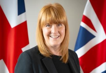 A head and shoulders photo of a woman with red hair and dark clothes standing in front of two Union Jack flags