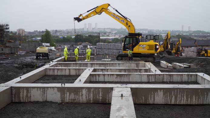 A yellow excavator lifts a concrete beam while men in PPE look on