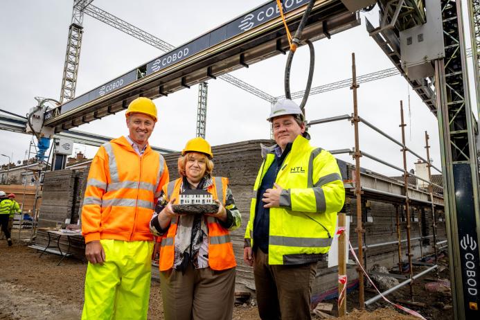 Three people in PPE on a construction site with one holding a model of a house