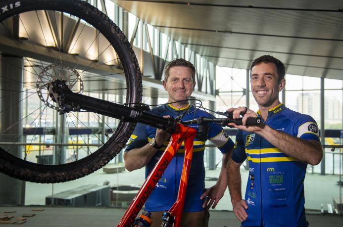 Two men in blue cycling clothes standing next to an upturned red bike and black wheel