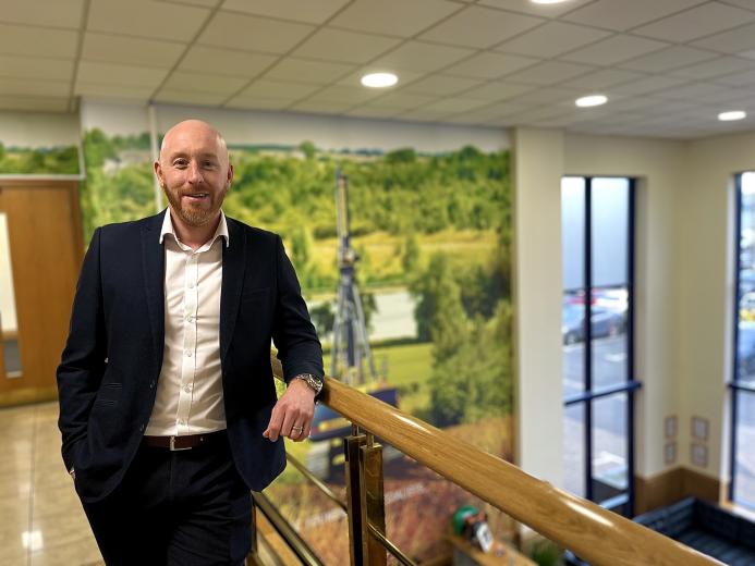 A bald man with a beard in an open necked shirt and dark suit standing on an internal office landing