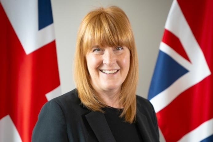 A head and shoulders photo of a woman with red hair and dark clothes standing in front of two Union Jack flags