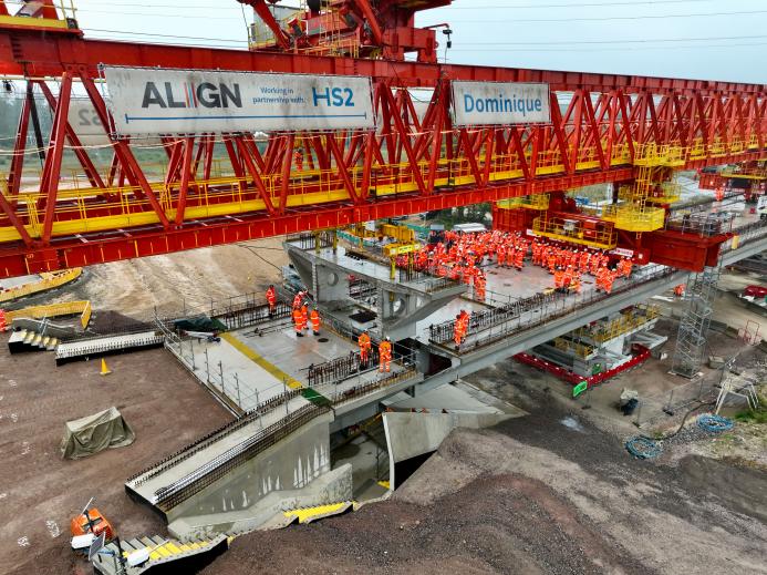 Construction workers in orange PPE install a precast concrete segment on a bridge deck using a bridge girder launcher