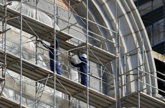 Two men in blue overalls and white hard hats on a scaffold cleaning a building