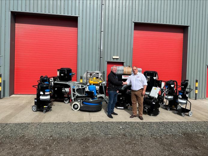 Two men shake hands infront of a range of floor grinding equipment, which is placed outside a grey warehouse with two, big red doors