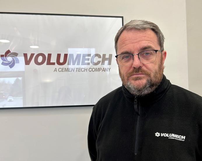 A man with grey hair, beard and glasses in a black fleece in front of a sign on the wall in an office