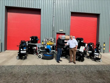 Two men shake hands infront of a range of floor grinding equipment, which is placed outside a grey warehouse with two, big red doors