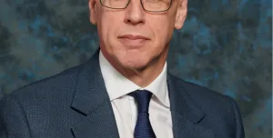 A studio head and shoulders photo of a man wearing glasses and a blue suit and tie