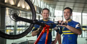 Two men in blue cycling clothes standing next to an upturned red bike and black wheel