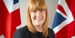 A head and shoulders photo of a woman with red hair and dark clothes standing in front of two Union Jack flags