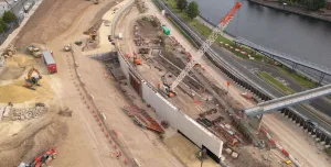 A large construction site by a river, including an orange crawler crane and other construction equipment