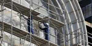 Two men in blue overalls and white hard hats on a scaffold cleaning a building