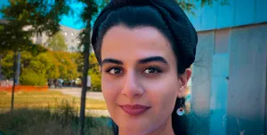 Head and shoulders picture of a young woman with dark hair, white jumper and necklace outside with trees in the background