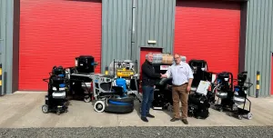 Two men shake hands infront of a range of floor grinding equipment, which is placed outside a grey warehouse with two, big red doors