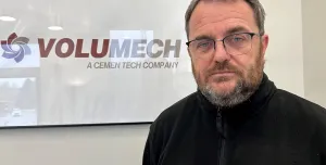 A man with grey hair, beard and glasses in a black fleece in front of a sign on the wall in an office