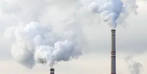 The top of two tall chimneys against a grey sky billow "smoke" into the atmosphere