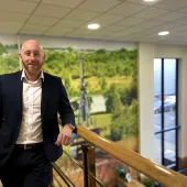 A bald man with a beard in an open necked shirt and dark suit standing on an internal office landing