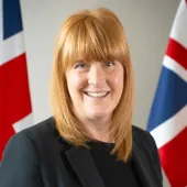 A head and shoulders photo of a woman with red hair and dark clothes standing in front of two Union Jack flags