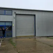 A bald man dressed in black outside a non-descript industrial estate