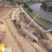 A large construction site by a river, including an orange crawler crane and other construction equipment