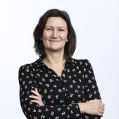 A head and shoulders photo of a woman with dark, straight, shoulder length hair, standing with arms folded wearing a black dress with small flowers printed on it