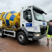 A man in yellow PPE stands in front of a white truck fitted with a yellow and blue concrete mixer