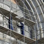 Two men in blue overalls and white hard hats on a scaffold cleaning a building