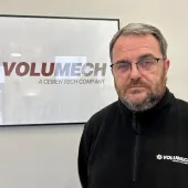 A man with grey hair, beard and glasses in a black fleece in front of a sign on the wall in an office