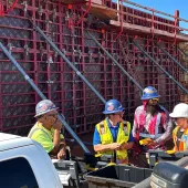 A group of construction workers in various colours of PPE stand in front of a wall of formwork