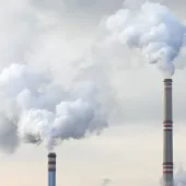 The top of two tall chimneys against a grey sky billow "smoke" into the atmosphere