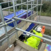 Two people in yellow PPE carry out a bridge inspection from a scaffold suspended from the side of the bridge