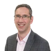 A head and shoulders shot of Peter Reekie of the Scottish Futures Trust will speak on sustainable infrastructure at the Glasgow Industry Day wearing glasses, an open necked pick shirt and a grey over-check jacket