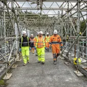 A group of construction workers walk along a concrete walkway, through a "tunnel" of scaffolding, wearing a variety of PPE , including orange hi vis jackets, yellow trousers, white hard hats and eye protectors