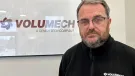 A man with grey hair, beard and glasses in a black fleece in front of a sign on the wall in an office