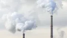 The top of two tall chimneys against a grey sky billow "smoke" into the atmosphere